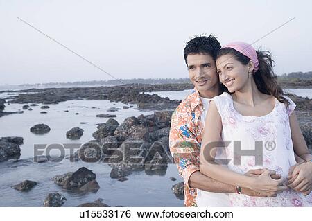 Stock Photograph - Young man embracing a young woman from behind on the coast. Fotosearch