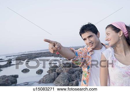Stock Image - Young man standing with a young woman on the coast and pointing forward. Fotosearch