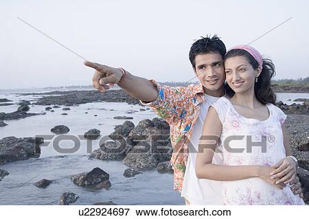 Stock Photo - Young man standing with a young woman on the coast and pointing forward. Fotosearch