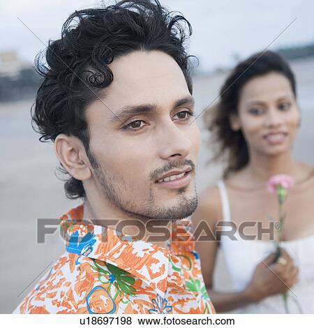 Close-up of a young man smiling with a young woman holding a flower behind him View Large Photo Image Stock Photo - Close-up of a young man smiling with a young woman holding a flower behind him. Fotosearch