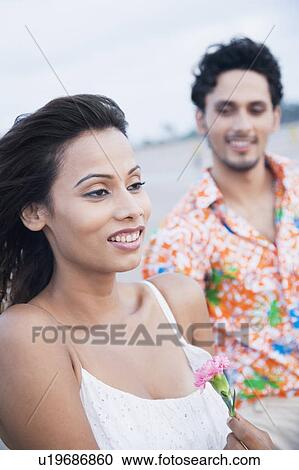 Close-up of a young woman holding a flower and smiling with a young man standing behind her View Large Photo Image Stock Image - Close-up of a young woman holding a flower and smiling with a young man standing behind her. Fotosearch