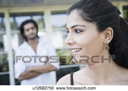 Close-up of a young woman smiling with a young man standing in the background View Large Photo Image Stock Image - Close-up of a young woman smiling with a young man standing in the background. Fotosearch