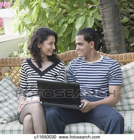 Young couple sitting with a laptop on a couch and looking at each other View Large Photo Image Stock Image - Young couple sitting with a laptop on a couch and looking at each other. Fotosearch