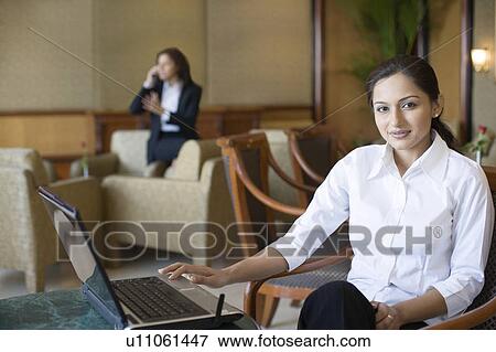 Stock Photo - Portrait of a businesswoman using a laptop with another businesswoman talking on a mobile phone in the background. Fotosearch