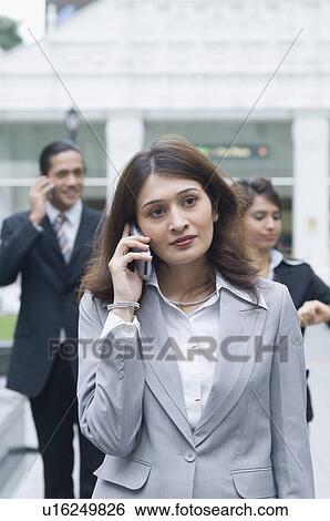 Businesswoman talking on a mobile phone with two business executives standing in the background View Large Photo Image Stock Photograph - Businesswoman talking on a mobile phone with two business executives standing in the background. Fotosearch