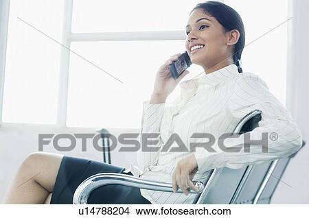 Close-up of a businesswoman sitting on a chair at an airport and talking on a mobile phone View Large Photo Image Picture - Close-up of a businesswoman sitting on a chair at an airport and talking on a mobile phone. Fotosearch
