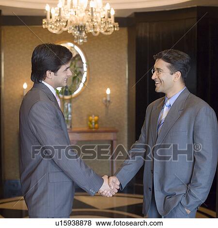 Stock Photo - Side profile of two businessmen shaking hands and smiling in a lobby. Fotosearch