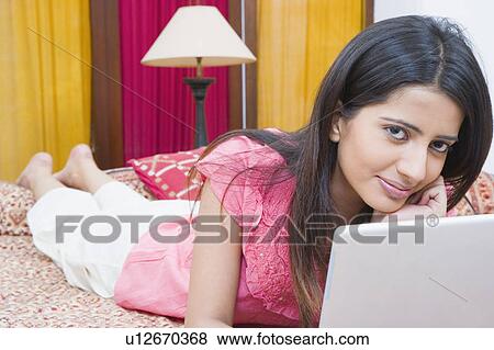 Stock Photo - Portrait of a young woman lying on the bed and using a laptop. Fotosearch