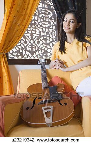 Stock Image - Young woman sitting on the arm of an armchair with a guitar. Fotosearch