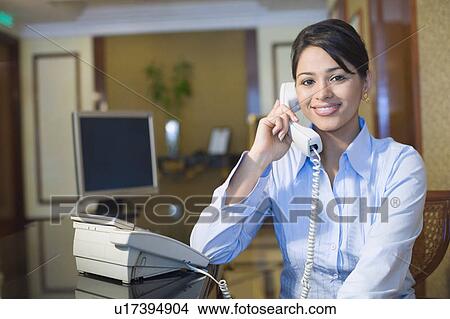 Picture - Portrait of a female receptionist sitting at a hotel reception and talking on the telephone. Fotosearch