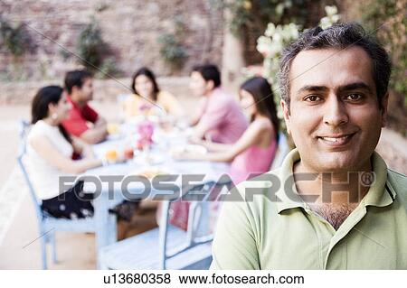 Portrait of a mid adult man smiling with his friends sitting at a dining table in the background View Large Photo Image Stock Photo - Portrait of a mid adult man smiling with his friends sitting at a dining table in the background. Fotosearch