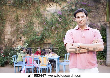 Portrait of a young man standing and his friends sitting at a dining table in the background View Large Photo Image Stock Photo - Portrait of a young man standing and his friends sitting at a dining table in the background. Fotosearch