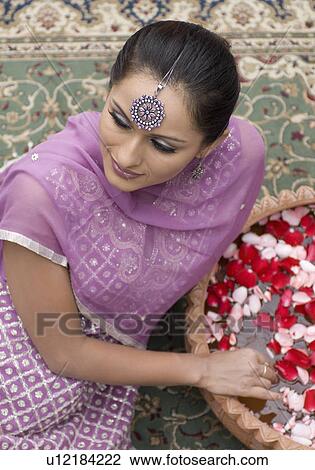 Stock Image - High angle view of a mid adult woman sitting on a carpet in salwar kameez with a bowl of petals. Fotosearch