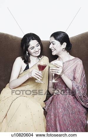 Picture - Two young women sitting on a couch toasting with martini glasses. Fotosearch