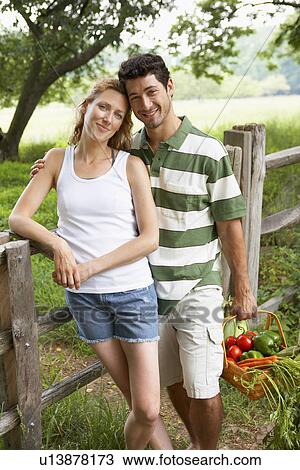 Stock Image - Husband and wife with vegetable basket standing by fence (portrait). Fotosearch