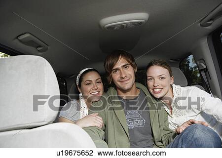 Stock Image - Young man sitting with two women on back seat (portrait). Fotosearch