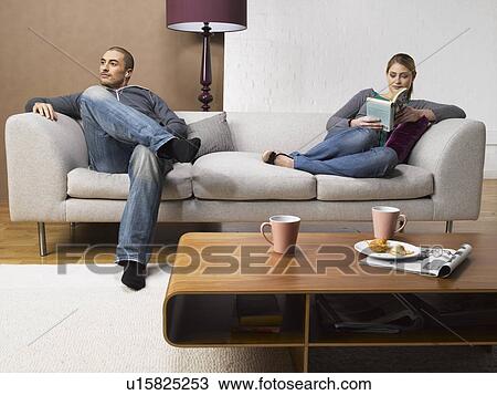 Stock Image - Young woman reading book and young man listening to MP3 player on sofa. Fotosearch