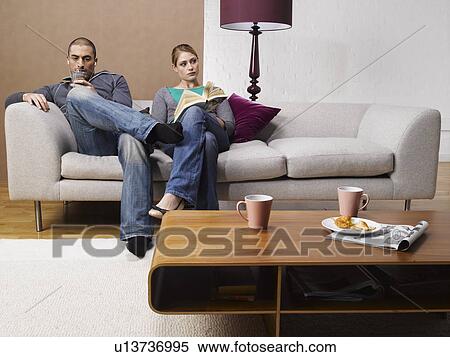 Stock Photography - Young woman reading book and young man listening to MP3 player on sofa. Fotosearch