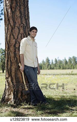 Stock Photo - Young man with axe leaning on tree. Fotosearch