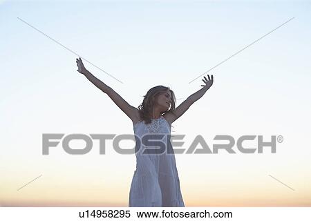 Stock Photography - Young woman in white dress raising hands at sunset. Fotosearch
