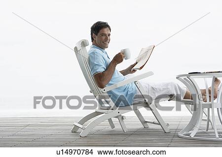 Picture - Mid adult man reading newspaper on deck near beach. Fotosearch