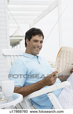 Stock Image - Mid adult man reading newspaper on lounge chair. Fotosearch