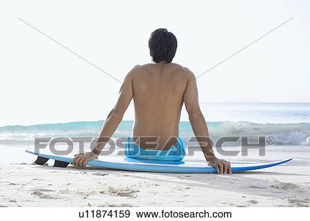 Stock Photo - Young man sitting on surfboard on beach, St. John, US Virgin Islands, USA. Fotosearch