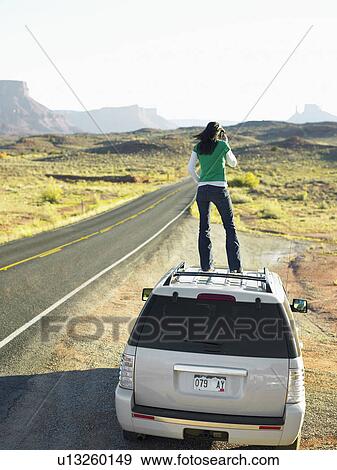 Stock Photo - Woman using cell phone on roof of car, rear view, Moab, Utah, USA. Fotosearch