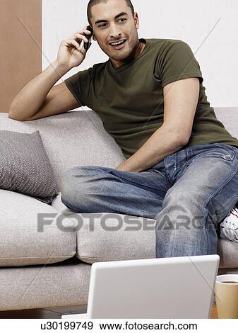 Stock Photo - Young man on cell phone on sofa, laptop on coffee table. Fotosearch
