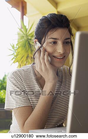 Stock Image - Young woman on cell phone and using laptop. Fotosearch