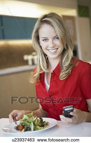 Stock Image - Young woman using cell phone while having lunch. Fotosearch
