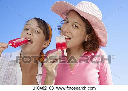 Stock Image - Two young women eating ice lollies (low angle view). Fotosearch
