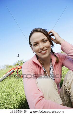 Picture - Young woman sitting on lawn, bicycle in background (portrait). Fotosearch