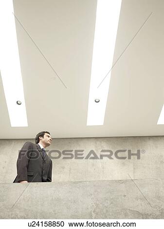 Stock Image - Businessman walking under strip lights (low angle view). Fotosearch