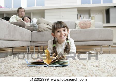 Stock Photo - Girl reading book on floor, parents on sofa in background. Fotosearch