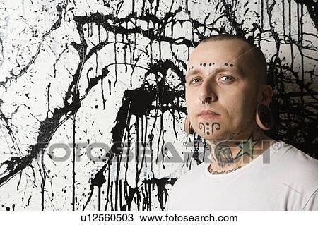 Stock Image - Tattooed and pierced man against paint splattered background. Fotosearch
