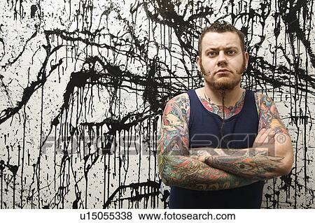 Stock Photo - Tattooed man with braided beard standing against paint splattered background. Fotosearch