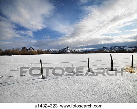 Stock Image - Barbed wire fence with snow covered ground and mountains in background. Fotosearch