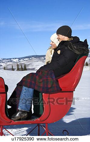 Stock Image - Side view of happy young couple seated in sleigh.. Fotosearch