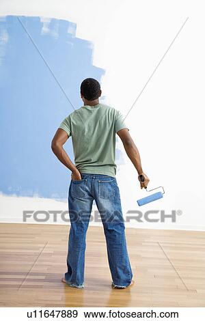 Stock Photo - African American young man standing next to half-painted wall with paint roller.. Fotosearch