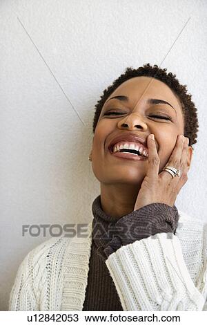 Stock Image - Close up head and shoulder of African-American woman standing against white wall smiling with hand on face and eyes closed.. Fotosearch