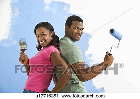 Stock Image - Couple next to half-painted wall as they stand back to back with paint utensils. Fotosearch