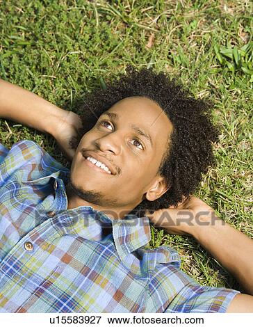 Stock Photo - Portrait of man lying in grass smiling with hands behind head.. Fotosearch