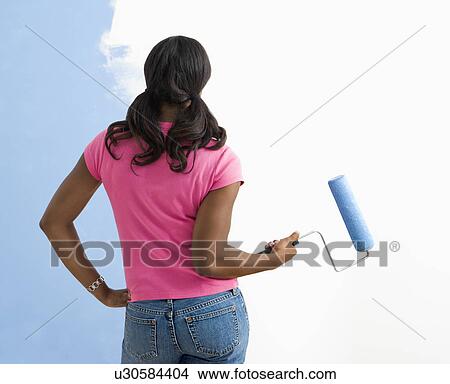 Picture - Woman standing next to half-painted wall with paint roller. Fotosearch