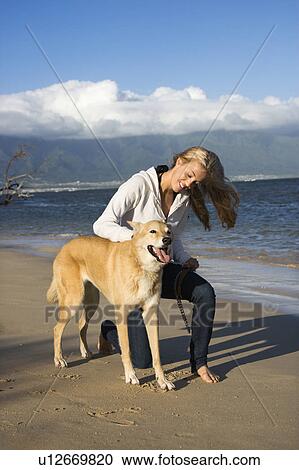 Stock Image - Caucasian woman petting brown dog on leash on Maui, Hawaii beach.. Fotosearch