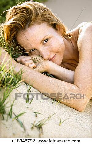 Stock Image - Close up portrait of attractive redheaded woman lying in grassy sand looking at viewer.. Fotosearch