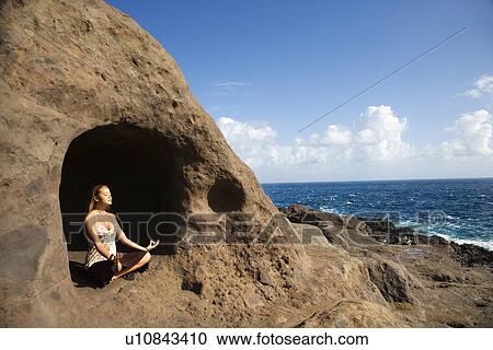 Stock Image - Young Asian woman mediatating in cave with coastline . Fotosearch