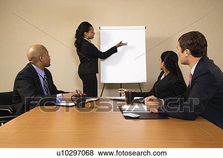 Stock Photo - Businesspeople sitting at conference table while businesswoman gives presentation.. Fotosearch