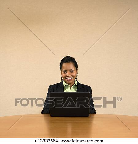 Picture - Businesswoman sitting at conference table smiling and typing on laptop computer.. Fotosearch
