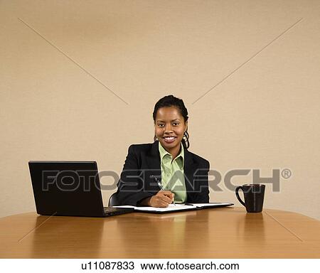 Stock Image - Businesswoman sitting at conference table with laptop computer and coffee cup smiling and writing in book. Fotosearch